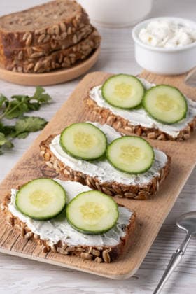 Slices of whole grain bread topped with cream cheese and fresh cucumber arranged beautifully at a rustic wooden table Three slices of nutritious whole grain bread are spread with creamy cheese and topped with fresh cucumber rounds, arranged on a wooden board for a light snack., 62897888.jpg RECORD DATE NOT STATED