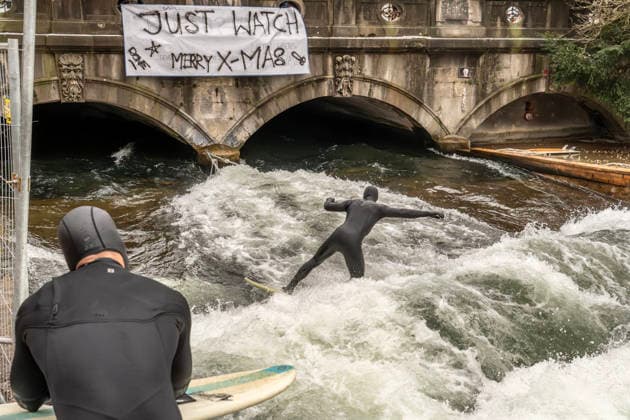 Eisbachwelle ist an Weihnachten zurück, ein Weihnachtswunder, seit Wochen war Stillstand, nun haben sich die Surfer selbst geholfen, provisorisch ein Brett ins Wasser gehängt, die Surfer sind glücklich, München, 26. Dezember 2025 Deutschland, München, 26. Dezember 2025, die Eisbachwelle ist zurück, seit gestern können wieder Surfer auf der Welle surfen, die berühmte Eisbachwelle wurde von den Surfern heimlich selber repariert, zwei Seile sind an der Brücke befestigt, dazwischen hängt ein Brett, das unter Wasser für den nötigen Widerstand sorgt und damit die neue alte Surfwelle aufbaut, Transparent an der Brücke: Just Watch, Merry X-mas , ein Weihnachtswunder, eine große Überraschung zu Weihnachten, seit fast zwei Monaten war