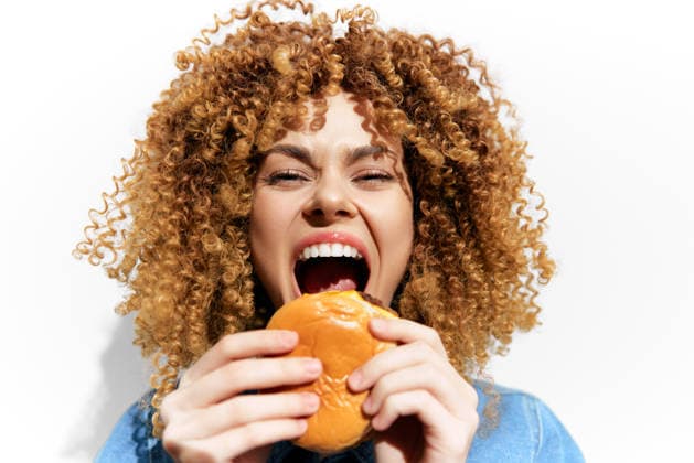 Cheerful young woman with curly hair joyfully biting into a delicious burger, captured on a clean white background, emphasizing vibrant colors and a fun food concept A vibrant and happy young woman with curly hair expresses joy while taking a big bite of a burger against a bright white backdrop, highlighting a playful food concept, model released, , 97807462.jpg RECORD DATE NOT STATED