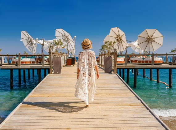 Junge Frau in einer weißen Spitzen-Tunika und einem Strohhut, die auf einem Holzsteg in Richtung eines luxuriösen Strandclubs mit Sonnenliegen und Sonnenschirmen geht. Young woman in a white lace tunic and straw hat walking along a wooden pier towards a luxury beach club with sun loungers and umbrellas for summer vacation and tropical resort concept (License=RF) ,model released, Symbolfoto Copyright: xZoonar.com AnnaxLuryex 24527504 ,model released, Symbolfoto ,property released