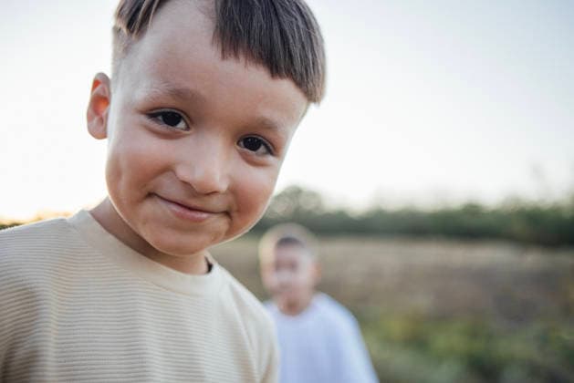 Ein kleiner Junge mit kurzen Haaren lächelt warm in die Kamera, umgeben von Natur, mit der verschwommenen Silhouette eines anderen Kindes im Hintergrund, und fängt einen fröhlichen Moment im Freien ein. Smiling young boy with short hair stands in a natural setting, with another child softly blurred in the background, creating a cheerful atmosphere of childhood joy (License=RF) ,model released, Symbolfoto Copyright: xZoonar.com DashaxPetrenkox 24547045 ,model released, Symbolfoto ,property released