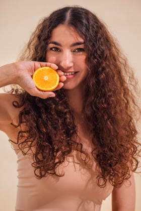 Junge Frau mit lockigem Haar hält eine frische Orangenscheibe vor ihr Gesicht und lächelt strahlend vor einem sanften beigen Hintergrund, wodurch sie einen gesunden Lebensstil und lebendige Energie vermittelt. Female with curly hair joyfully holds orange slice near her face, radiating happiness and health, set against a warm beige backdrop, emphasizing freshness and vitality (License=RF) ,model released, Symbolfoto Copyright: xZoonar.com DashaxPetrenkox 24582731 ,model released, Symbolfoto ,property released