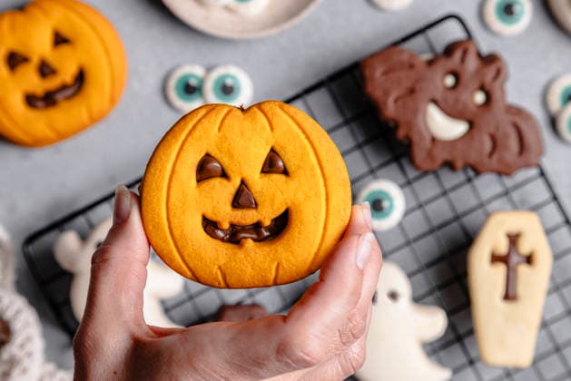Halloween cookies with pumpkin and bat designs Top view of a cropped unrecognizable person holding a pumpkin-shaped Halloween cookie. Background features bat and ghost cookies on a cooling rack over a gray surface. Model Released M-aCreativeStudio_ID32786_1116120_010 Copyright: xMxAxCreativexStudiox