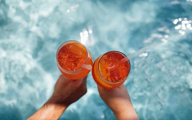 Zwei Hände halten Gläser mit orangefarbenen Cocktails mit Eiswürfeln, die teilweise in einem klaren Swimmingpool eingetaucht sind, Sonnenlicht reflektiert auf der Wasseroberfläche. Two hands grasp glasses of aperol spritz filled with orange cocktails and ice, partially submerged in a clear pool, with sunlight creating reflections on the water (License=RF) Copyright: xZoonar.com DashaxPetrenkox 24646524
