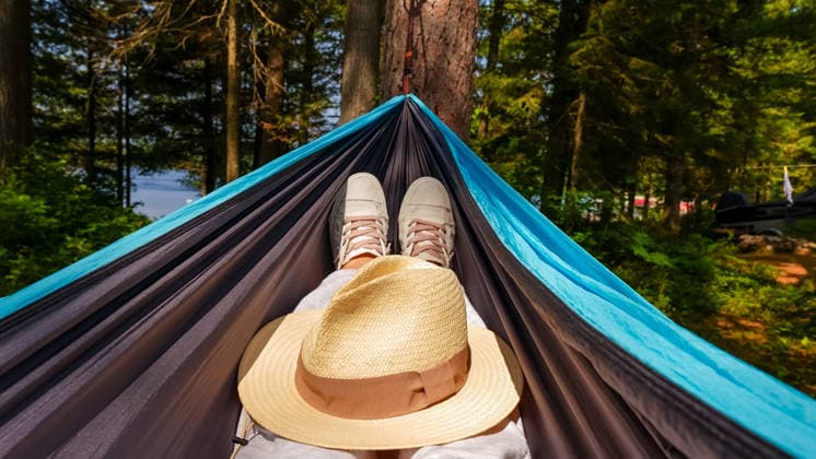 Person, die sich in einer Hängematte entspannt, Turnschuhe und einen Strohhut trägt und von üppig grünen Bäumen in der Nähe eines ruhigen Sees umgeben ist. Individual resting in a hammock with sneakers and a straw hat, nestled among vibrant green trees beside a calm lake (License=RF) Copyright: xZoonar.com IuliiaxDombrovskaiax 24639724