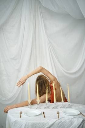 Man sieht eine Frau, die sich anmutig an einem mit Kerzen gedeckten Tisch beugt. Der Tisch hat weiße Teller und Gabeln, und der Hintergrund besteht aus weichem weißem Stoff. Es ist Abend und die Szene spielt drinnen A woman is seen bending gracefully at a table set with candles. The table has white plates and forks, and the backdrop features soft white fabric. It is evening and the scene is set indoors Copyright: imageBROKER AlekseixIsachenko ibxaii16668431.jpg ,model released, Symbolfoto ,property released