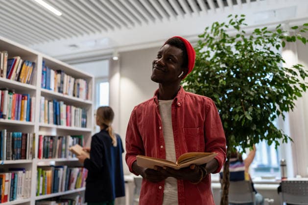 Ein fröhlicher schwarzer Student steht mit einem Buch in der Universitätsbibliothek und denkt über einen guten Job nach dem Abschluss nach. Satisfied African American student stands in library with open book looks dream about passed exam enjoy interesting effective studies in higher school. Successful excellent guy learner get scholarship (License=RF) ,model released, Symbolfoto Copyright: xZoonar.com DmitriixMarchenkox xEnvatox 24684615 ,model released, Symbolfoto ,property released