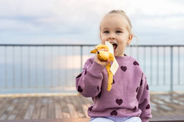 Toddler enjoying a banana on outdoor deck A happy toddler girl wearing a purple sweater with heart designs enjoys eating a banana while sitting on an outdoor deck. The scene shows a serene background of the sky and sea. Model Released AlinaKandybka_ID33645_1171225_026 Copyright: xAlinaxKandybkax RECORD DATE NOT STATED