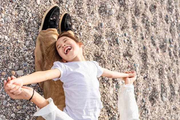 Daughter laughing joyfully on a pebbly beach A joyful daughter laughs while lying on a pebbly beach, holding hands with a parent. Her carefree spirit and happiness are captured in this sunny, heartwarming moment. Model Released AlinaKandybka_ID33946_1195165_005 Copyright: xAlinaxKandybkax RECORD DATE NOT STATED