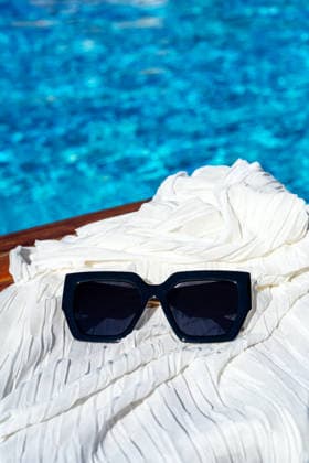 Stylish sunglasses on fabric with refreshing pool backdrop A pair of sleek black sunglasses rests on white fabric, set against a glistening blue pool backdrop. This stylish setup captures the essence of sunny relaxation and leisure. Model Released IrinaChetverikova_ID33496_1166281_001 Copyright: xIrinaxChetverikovax RECORD DATE NOT STATED