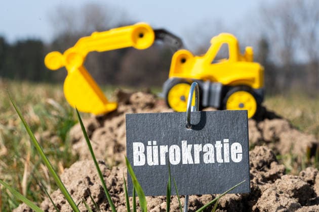 Bavaria, Germany, March 6, 2026: PHOTOMONTAGE, A toy excavator stands on a small construction site next to a sign reading Bureaucracy.? The image symbolizes administrative hurdles, permits, and regulations that can delay construction projects and complicate construction planning. *** FOTOMONTAGE, Ein Spielzeugbagger steht auf einer kleinen Baustelle neben einem Schild mit der Aufschrift Bürokratie. Das Bild symbolisiert administrative Hürden, Genehmigungen und Vorschriften, die Bauprojekte verzögern und Bauplanung erschweren können.