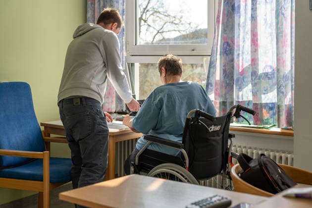 Pfleger unterstützt einen Patienten im Rollstuhl bei einer Aufgabe am Tisch in einer Pflegeeinrichtung. Die Szene zeigt den Alltag in der Pflege, in dem Betreuung, Unterstützung und menschliche Fürsorge eine zentrale Rolle spielen.