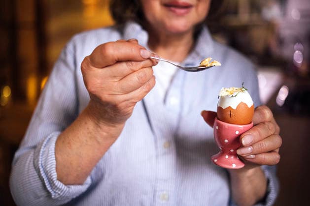 IMAGO Creative, Ostern, Bräuche, Osterbrunch Eine ältere Frau in einem blau gestreiften Hemd hält einen Löffel mit einem weichgekochten Ei in einem rosa Eierbecher in der Hand; Küchenszene mit warmer Beleuchtung und unscharfen Hintergrundelementen Senior woman in blue striped shirt prepares to eat soft-boiled egg from pink egg cup, with kitchen elements softly blurred in the background (License=RF) ,model released, Symbolfoto Copyright: xZoonar.com DashaxPetrenkox 24823835 ,model released, Symbolfoto ,property released