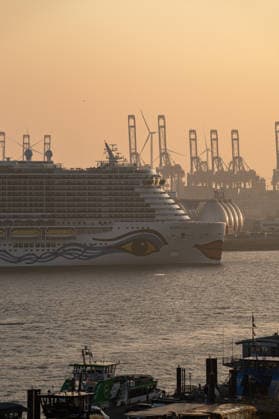 Deutschland, Hamburg: Kreuzfahrtschiff AIDAnova auf der Elbe vor Containerterminal und Industrieanlagen, Teil der Flotte von AIDA Cruises, markanter AIDA-Kussmund am Bug im Gegenlicht, im Hintergrund Hafenkräne und große kugelförmige Speicheranlagen, Windkraftanlagen als Zeichen der Energiewende, Vordergrund mit kleineren Hafen- und Versorgungsschiffen, dichter industrieller Kontext des Hamburger Hafens, einer der größten Seehäfen Europas, Kontrast zwischen Kreuzfahrttourismus, Energieinfrastruktur und Schwerindustrie, warmes Abendlicht mit Dunst und Silhouettenwirkung, maritime Wirtschaft und Logistik, dokumentarische Stadt- und Industriefotografie. Hamburg *** Germany, Hamburg cruise ship AIDAnova on the Elbe in front of containe Copyright: xStefanxTrochax