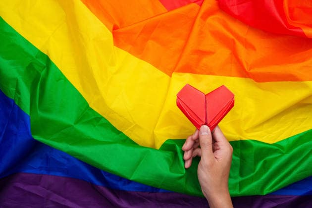 Hand, die ein rotes Origami-Herz auf einer Regenbogenfahne hält A hand holding a red origami heart placed on a vibrant rainbow flag, symbolizing love, pride, and support for the LGBTQ community. (License=RF) Copyright: xZoonar.com WORAWEExMEEPIANx 24850258