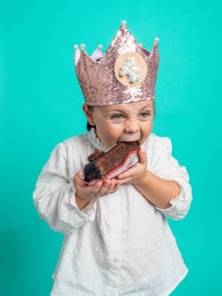 Little girl celebrates with a chocolate treat A young girl joyfully bites into a chocolate treat while wearing a sparkling crown and a white dress, celebrating her birthday in a vibrant studio setting. Model Released MaciaPuiggros_ID34290_1237089_005 Copyright: xx RECORD DATE NOT STATED