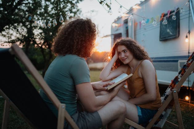 Couple having romantic camping trip in nature. Man reading book to girlfriend. Caravan traveling for young people. Couple having romantic camping trip in nature. Man reading book to girlfriend. Caravan traveling for young people model released