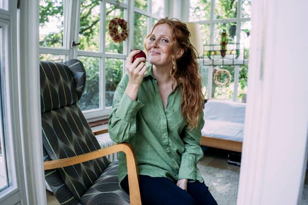 Smiling woman with long hair holding apple and sitting on chair at home model released, Symbolfoto property released, JOSEF26304