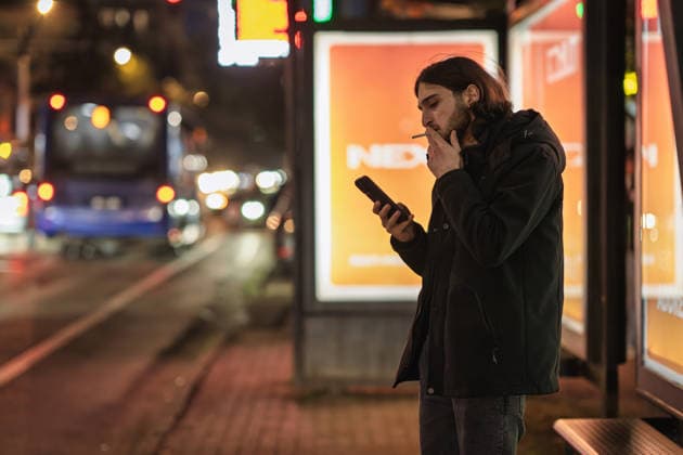 Batumi, georgia 21 april 2026: young man standing at a bus stop at night, smoking a cigarette, and using his mobile phone with city lights in background Man smoking cigarette using smartphone at night bus stop in batumi Copyright: xx Batumi Edit 1106