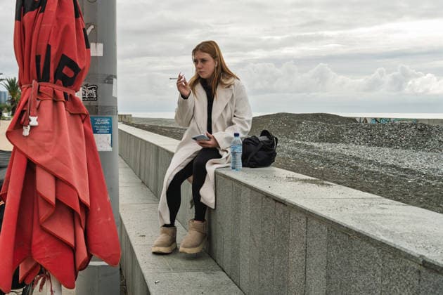 Batumi, georgia 21 april 2026: young woman in trench coat sits on seaside wall in batumi, smoking and checking her phone on an overcast promenade stroll Woman smoking cigarette on batumi beach promenade Copyright: xx Batumi Edit 1220