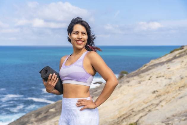 Frau lächelt, hält eine gerollte Yogamatte auf einer felsigen Klippe und blickt auf das strahlend blaue Meer, was für einen gesunden Lebensstil, Fitness und Wellness steht Woman smiling, holding a rolled yoga mat on a rocky cliff, looking towards the bright blue ocean, representing a healthy lifestyle, fitness, and wellness journey Copyright: imageBROKER UnaixHuizi ibxuhu16618242.jpg ,model released, Symbolfoto ,property released