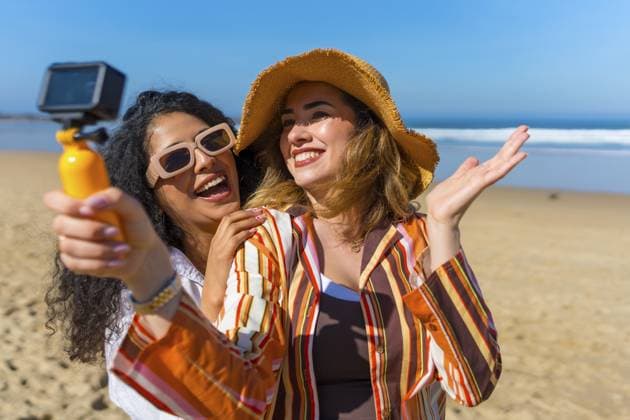 Zwei lächelnde junge Frauen, die mit einer kleinen Videokamera an einem sonnigen Sandstrand Inhalte erstellen, Spaß haben und gemeinsam den Sommerurlaub am Meer genießen Two smiling young women creating content with a small video camera on a sunny sandy beach, having fun and enjoying summer holiday together by the ocean Copyright: imageBROKER UnaixHuizi ibxuhu16975588.jpg ,model released, Symbolfoto ,property released