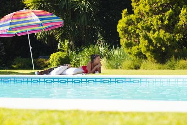 Adult woman lying on stomach at backyard pool edge wearing red swimsuit under striped umbrella, model released, property released, 19.03.2025 06:00:00, 12839384 Copyright: xIMS-260409_1xcreatorid:x372x 12839384