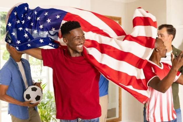 Diverse male fans wearing red tee, jersey cheering, draping US flag, holding soccer ball at home, model released, property released, Cape Town, South Africa, 18.03.2026 06:00:00, 12882234 Copyright: xIS-260415_1xcreatorid:x393x 12882234