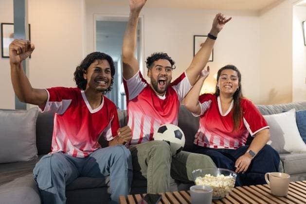 Friends wearing red jerseys sitting at home cheering holding soccer ball popcorn mugs smartphone, model released, property released, Cape Town, South Africa, 26.08.2025 06:00:00, 8921133 Copyright: xIS-251205xcreatorid:x393x 8921133