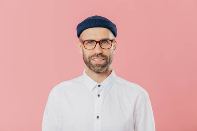 Bärtiger Mann mit blauer Mütze und Brille, der in einem weißen Hemd vor einem rosa Hintergrund in die Kamera lächelt Bearded Man in Blue Beanie Hat and Glasses Smiling at Camera in White Shirt Against Pink Background (License=RF) ,model released, Symbolfoto Copyright: xZoonar.com ViorelxKurnosovx 25104028 ,model released, Symbolfoto ,property released