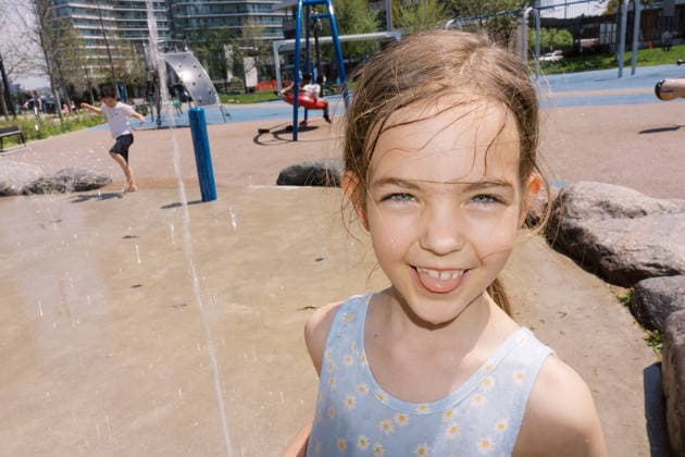Lächelndes Mädchen genießt den städtischen Wasserspielplatz A young girl smiles at the camera while playing at a splash pad in a city park, with water jets spraying and other children playing in the background. (License=RF) ,model released, Symbolfoto Copyright: xZoonar.com VolodymyrxNadtochiix 25118168 ,model released, Symbolfoto ,property released