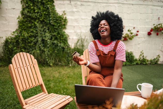 Eine junge Frau lacht beim Online-Shopping mit Kreditkarte im Freien Young adult woman enjoying online shopping, laughing while holding a credit card and using a laptop in a garden (License=RF) ,model released, Symbolfoto Copyright: xZoonar.com JelenaxCvetkovicx 25122395 ,model released, Symbolfoto ,property released