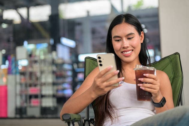 IMAGO Creative, Food, Trend, Iced Coffee Eine lächelnde Thailänderin, die im Freien ihr Smartphone benutzt, während sie einen Eiskaffee in der Hand hält. Horizontal medium shot of a happy young Isan Thai female customer looking down and smiling while operating her smartphone held in both hands. Seated relaxed in a green folding camping chair outdoors, she concurrently holds a clear plastic takeaway cup of iced coffee or iced tea, featured in a wide composition with excellent copy space on the left side. (License=RF) ,model released, Symbolfoto Copyright: xZoonar.com TonixRantalax 25138614 ,model released, Symbolfoto ,property released