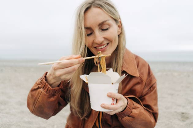 Eine blonde Frau isst draußen mit Stäbchen asiatische Fertignudeln aus einer Schachtel. Blonde woman eating Asian takeout noodles with chopsticks from a box outdoors. (License=RF) ,model released, Symbolfoto Copyright: xZoonar.com TITOVAxILONAx 25185296 ,model released, Symbolfoto ,property released