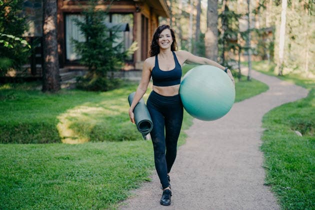 Eine lächelnde Frau trägt einen Gymnastikball und eine Trainingsmatte und geht auf einem Weg in der Nähe eines Holzhauses im Wald spazieren Smiling woman carries fitness ball and exercise mat walking along path near wooden house in forest (License=RF) ,model released, Symbolfoto Copyright: xZoonar.com ViorelxKurnosovx 25185751 ,model released, Symbolfoto ,property released