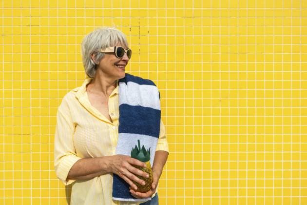 Seniorin mit grauen Haaren lächelt, trägt eine Sonnenbrille, hält ein gestreiftes Handtuch und eine ananasförmige Flasche gegen eine leuchtend gelb geflieste Wand und genießt die Sommerfreizeit Senior woman with gray hair smiling, wearing sunglasses, holding a striped towel and a pineapple shaped bottle against a bright yellow tiled wall, embracing summer leisure Copyright: imageBROKER UnaixHuizi ibxuhu17868376.jpg ,model released, Symbolfoto ,property released