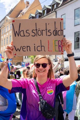 Augsburg, Bavaria, Germany - June 13, 2026: Participants carrying rainbow flags and wearing colorful costumes take part in Christopher Street Day (CSD) in Augsburg. The parade through the city center promotes the rights of LGBTQ+ people as well as social diversity and tolerance. A woman holds a homemade sign in rainbow colors that reads: WHAT™S IT TO YOU WHO I LOVE *** Teilnehmerinnen und Teilnehmer mit Regenbogenflaggen und bunten Kostümen nehmen am Christopher Street Day (CSD) in Augsburg teil. Der Demonstrationszug durch die Innenstadt wirbt für die Rechte von LGBTQ+-Menschen sowie für gesellschaftliche Vielfalt und Toleranz. Eine Frau hält ein selbst gebasteltes Schild mit Aufschirft: WAS STÖRTS DICH WEN ICH LIEBE in Regenbogenfa