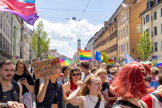 Augsburg, Bavaria, Germany June 13, 2026: Participants carrying rainbow flags and wearing colorful costumes take part in Christopher Street Day (CSD) in Augsburg. The parade through the city center promotes the rights of LGBTQ+ people as well as social diversity and tolerance. A participant holds a sign reading: QUEER WAS ALWAYS HERE *** Teilnehmerinnen und Teilnehmer mit Regenbogenflaggen und bunten Kostümen nehmen am Christopher Street Day (CSD) in Augsburg teil. Der Demonstrationszug durch die Innenstadt wirbt für die Rechte von LGBTQ+-Menschen sowie für gesellschaftliche Vielfalt und Toleranz. Teilnehmerin hält ein Schild: QUEER WAS ALWAYS HERE