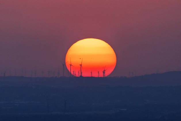 Sonnenuntergang in der Bonn Windräder eines Windparks zeichnen sich als Silhouette vor der riesigen glutroten Sonnenscheibe ab. Auf der Sonne sind Sonnenflecken zu erkennen. Der Hitze-Dunst nach einem Hitzetag mit bis zu 35 Grad lässt die Sonne beim Sonnenuntergang über dem Bonner Raum überdimensional groß erscheinen. Blick vom Petersberg im Siebengebirge. Königswinter Petersberg Nordrhein-Westfalen Deutschland *** Sunset in Bonn: The wind turbines of a wind farm stand out as a silhouette against the huge, glowing-red disk of the sun. Sunspots are visible on the sun. The heat haze following a hot day with temperatures reaching up to 35 degrees makes the sun appear enormous as it sets over the Bonn area. View from Petersberg in the Sie Copyright: xMarcxJohnx