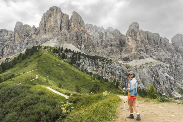 Wanderin mit Wanderstock, die sich dreht, um in die Kamera zu schauen, geht auf einem kurvenreichen Feldweg entlang des grünen Alpenkamms, majestätische zerklüftete Cir Gipfel im Hintergrund, Grödner Dolomiten, Italien Female hiker with trekking pole turning to look at camera walking winding dirt path along green alpine ridge majestic jagged Cir peaks background Passo Gardena Dolomites Italy Copyright: imageBROKER EvaldasxMikoliunas ibxemi18154186.jpg ,model released, Symbolfoto ,property released