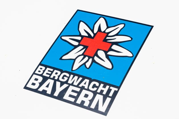 14. Landestag der Verkehrssicherheit, Nürnberg, 27.06.2026 Logo der Bergwacht Bayern mit stilisiertem Edelweiss und rotem Kreuz auf blauem Grund. Das Schild ist auf einem Fahrzeug oder Ausstattungsgegenstand angebracht. Nürnberg Bayern Deutschland *** 14th State Road Safety Day, Nuremberg, June 27, 2026 Logo of the Bavarian Mountain Rescue Service featuring a stylized edelweiss and a red cross on a blue background. The sign is affixed to a vehicle or piece of equipment. Nuremberg, Bavaria, Germany 20260627-286A0494-M4000