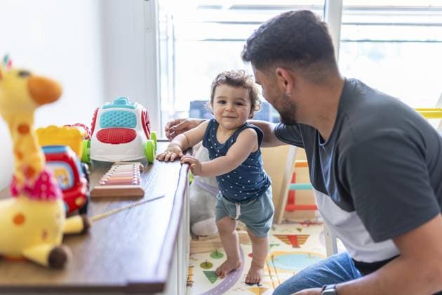 Lächelnder Junge, der sich an einem Schrank festhält, während er sich stabilisiert und während spielerischer Bindungen und ersten Schritten zu Hause zu seinem Vater schaut, freudiger Familienmoment des Lernens und Vertrauens Smiling baby boy holding onto a cabinet as he steadies himself and looks to his father during playful bonding and first steps at home, joyful family moment of learning and trust Copyright: imageBROKER UnaixHuizi ibxuhu18091862.jpg ,model released, Symbolfoto ,property released