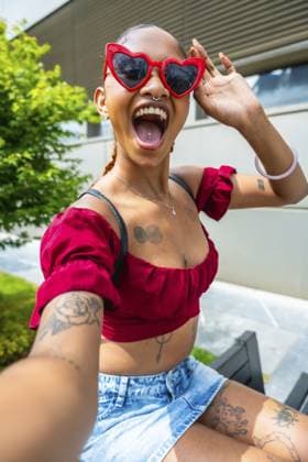Junge Frau mit roter Herz Sonnenbrille, Septum und Zungenpiercings und Körpertattoos, fotografiert ein Selfie und drückt Glück und Aufregung im Freien aus Young woman with red heart sunglasses, septum and tongue piercings, and body tattoos capturing a selfie, expressing happiness and excitement outdoors Copyright: imageBROKER UnaixHuizi ibxuhu18093798.jpg ,model released, Symbolfoto ,property released