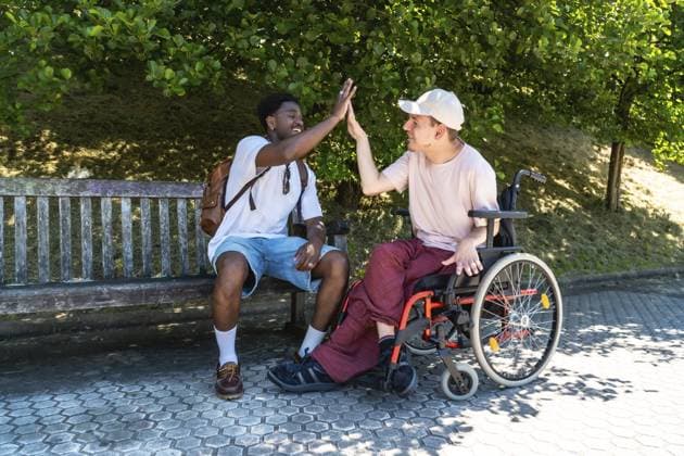 Zwei fröhliche männliche Freunde, einer im Rollstuhl, klatschen auf einer sonnigen Parkbank, umgeben von grünem Laub, um Inklusion, Unterstützung und Freundschaft zu feiern Two cheerful male friends, one in a wheelchair, share a high five on a sunny park bench surrounded by green foliage, celebrating inclusion, support and friendship Copyright: imageBROKER UnaixHuizi ibxuhu18194654.jpg ,model released, Symbolfoto ,property released