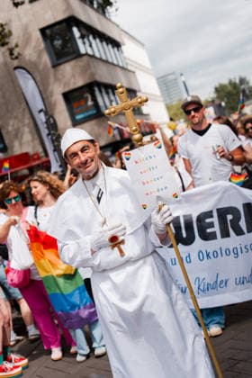 05.07.26, Cologne Pride 2026, parade and demonstration for diversity and equality A participant dressed as a pope holds a cross-shaped sign reading Kirche sollte bunter werden (The church should become more colorful) while walking with a queer church group in the parade. Cologne Cologne NRW Germany