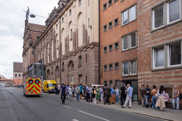 Feuerwehr-Großeinsatz am Nürnberger Opernhaus nach Brand in Tiefgarage, Nürnberg, 09.07.2026 Mitarbeiter und Besucher des Staatstheaters Nürnberg stehen nach der Evakuierung auf dem Gehsteig vor dem Gebäude. Auslöser war ein brennendes Auto in der angrenzenden Tiefgarage, das Feuer griff auf weitere Fahrzeuge über. Ein Rettungswagen der Feuerwehr steht auf der Straße vor Ort. Nürnberg Bayern Deutschland *** Large-Scale Fire Department Operation at the Nuremberg Opera House Following a Fire in the Underground Parking Garage, Nuremberg, July 9, 2026 Employees and visitors of the Nuremberg State Theater stand on the sidewalk in front of the building after the evacuation. The fire was triggered by a burning car in the adjacent undergr 20260709-286A1422-M4000