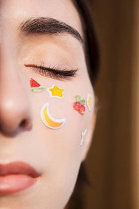 Nahaufnahme einer jungen Frau mit Aufklebern im Gesicht, die Obst- und Sternmotive zeigen und einen verspielten und kreativen Ausdruck von Schönheit in einer sanft beleuchteten Innenraumkulisse unterstreichen Young woman with closed eyes has colorful stickers on her face, including fruit and star shapes, in a softly lit indoor environment (License=RF) ,model released, Symbolfoto Copyright: xZoonar.com DashaxPetrenkox 25252005 ,model released, Symbolfoto ,property released