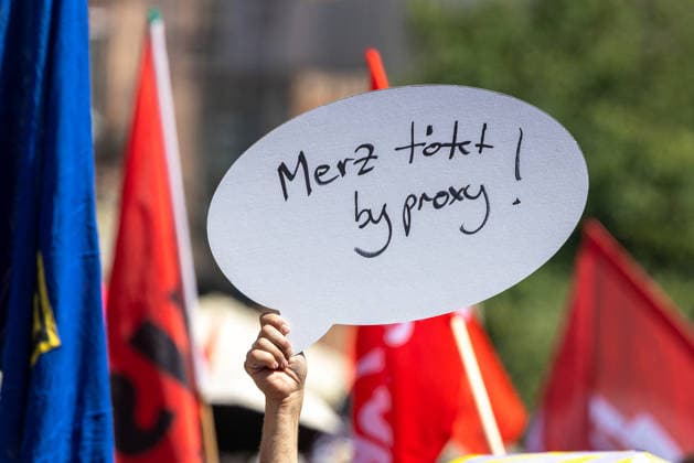 Demonstration für Solidarität statt Sozialabbau, Nürnberg, 11.07.2026 Eine Demonstrantin hält ein Schild in Sprechblasenform mit der Aufschrift Merz tötet by proxy! in die Höhe. Im Hintergrund sind rote und blaue Fahnen der Kundgebung zu sehen. Nürnberg Bayern Deutschland *** Demonstration for Solidarity Instead of Social Cuts, Nuremberg, July 11, 2026. A demonstrator holds aloft a speech bubble-shaped sign reading Merz kills by proxy. Red and blue rally flags can be seen in the background. Nuremberg, Bavaria, Germany 20260711-1V9A5220-M4000