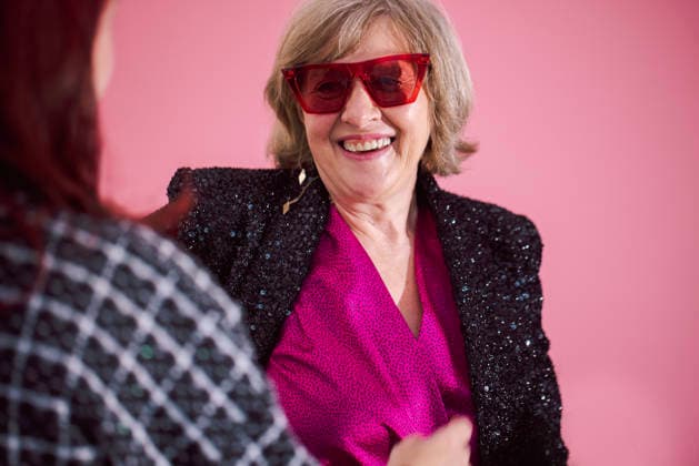 Eine ältere Frau mit einer stilvollen roten Sonnenbrille und einem glitzernden schwarzen Blazer lächelt, während sie sich vor einem rosa Hintergrund in einer farbenfrohen Innenraumkulisse mit einer anderen Person unterhält. Senior woman in red sunglasses and black blazer engages with another person in a lively indoor space featuring a pink backdrop (License=RF) ,model released, Symbolfoto Copyright: xZoonar.com DashaxPetrenkox 25282694 ,model released, Symbolfoto ,property released