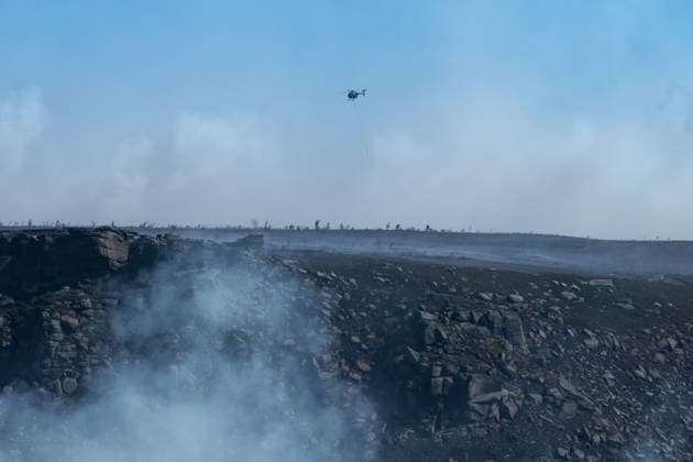 A helicopter carries water over smouldering moorland close to Dove Stone Reservoir in Greater Manchester on 13 July 2026. Greater Manchester Police have arrested a woman in connection with a large moorland fire at Dove Stone Reservoir in Greenfield. The blaze, burning since Saturday, is under active investigation, with officers following leads regarding reports of fireworks nearby. The 20-year-old suspect remains in custody as the cause of the fire is yet to be officially determined. Oldham Greater Manchester England Dovestone Reservoir  Copyright: xMattxWilkinsonx